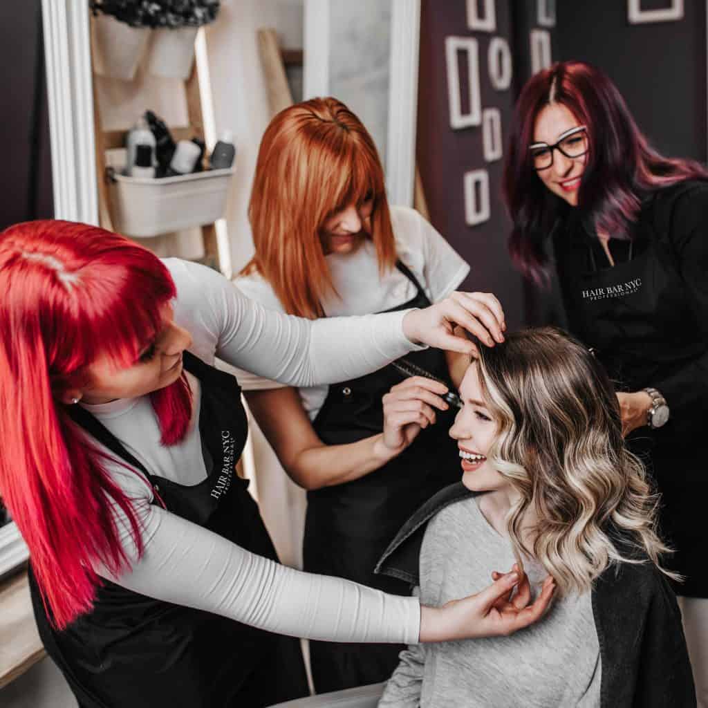 Three hair stylists with red and purple hair work on a smiling woman's hair in a salon. The woman is seated and wearing a black cape.