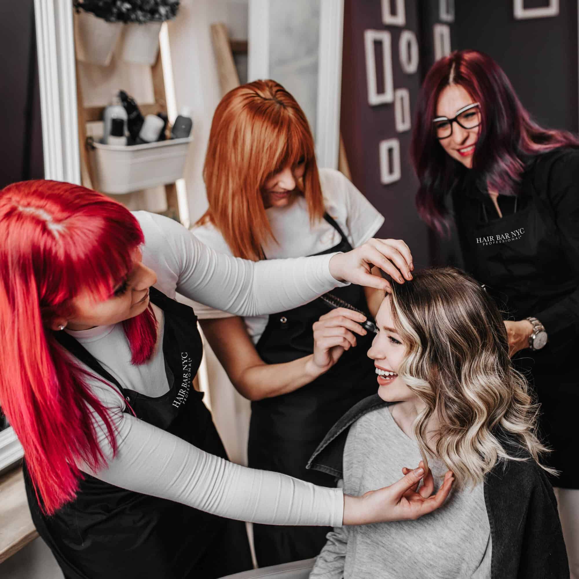 Three hair stylists with red and purple hair work on a smiling woman's hair in a salon. The woman is seated and wearing a black cape.