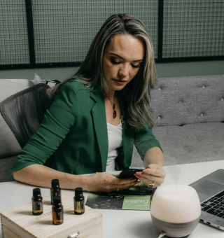 A woman in a green blazer sits at a desk looking at her phone, with essential oil bottles, a diffuser, and a laptop in front of her.