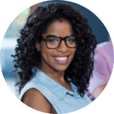 Woman with curly hair and glasses smiling at the camera, wearing a sleeveless denim shirt, indoors with a blurred background.