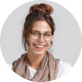 A young woman with brown hair in a bun, wearing glasses, a light scarf, and a white top, smiling at the camera against a plain background.