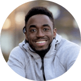 A man with short hair and a beard, wearing a light gray hoodie, smiles while looking at the camera outdoors.