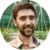 A man with short brown hair and a beard, wearing a checked shirt, is smiling in a greenhouse with hanging plants in the background.