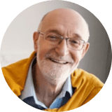 Older man with glasses and a beard, smiling, wearing a yellow sweater over a collared shirt, indoors with a neutral background.