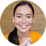 A woman with long dark hair, wearing a yellow shirt, smiles at the camera with hands gently clasped under her chin against a blurred brown background.