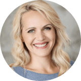 A woman with light blonde wavy hair and blue eyes is smiling at the camera, wearing a light blue top. The background is gray and blurred.