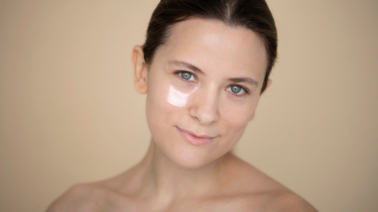 A person with light skin and brown hair wears under-eye patches and looks at the camera against a beige background.
