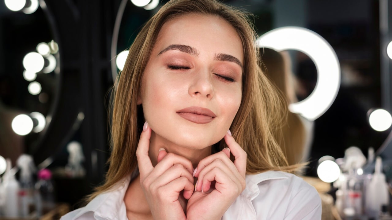 Woman with closed eyes and neutral expression touching her face with both hands, in a well-lit room with mirrors and round lights in the background.