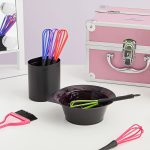 The Color Dye Mixer set, featuring colorful plastic whisks, a brush, a mixing bowl with dye, and a pink storage box, is displayed on a white table near a mirror.
