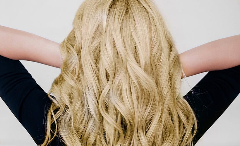 Person with long, wavy blonde hair seen from behind, wearing a dark top with arms raised, hands touching the back of their head.
