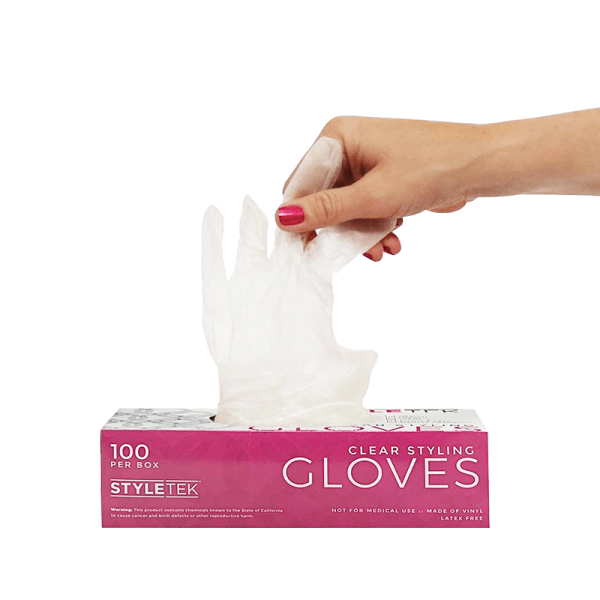 A hand pulls a clear disposable glove from a pink and white box labeled "Clear Deluxe Vinyl Gloves – Powder Free, 100 gloves per box.