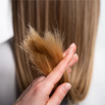A hand holds the dry, split ends of long, straight, light brown hair, showing damage that G&C Cream Developer 40 Volume can help address.
