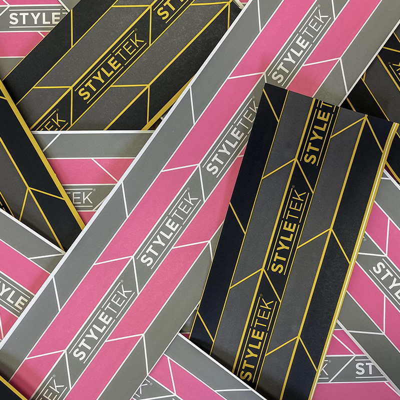 Close-up of multiple overlapping Pop Up Foil 5 x 11 - Heavy Emboss (500CT) packages in pink, gray, black, and gold geometric STYLETEX designs.