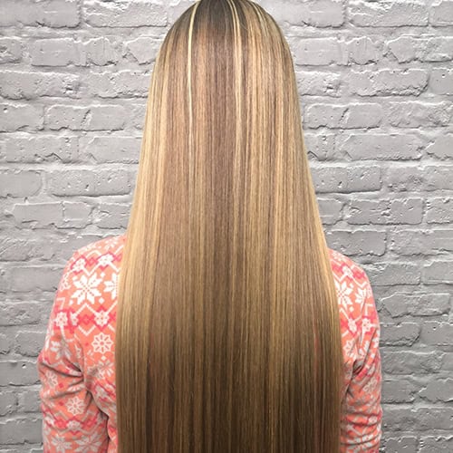 Person with long, straight, blonde-highlighted hair, wearing a pink and white patterned sweater, standing in front of a light gray brick wall.