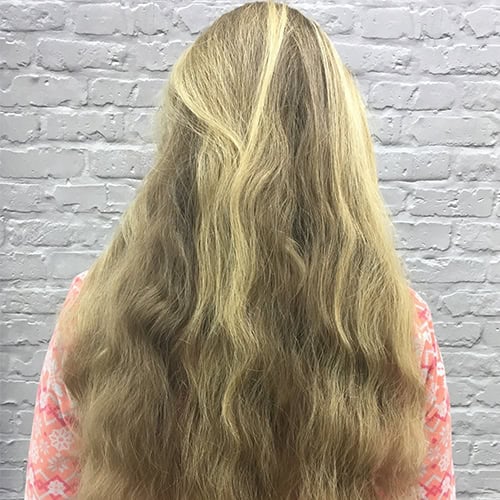 A person with long, wavy, blonde hair shown from the back, standing in front of a light gray brick wall.