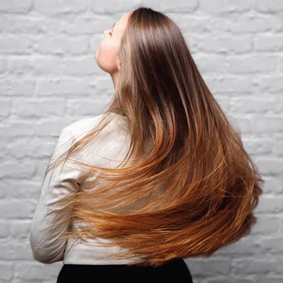 Person with long, straight, light brown hair wearing a light-colored top, shown from the back in front of a white brick wall.