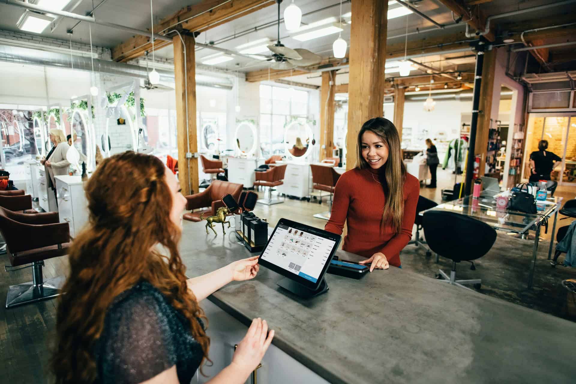 Two women interact at a salon reception desk; one is behind the counter using a point-of-sale system, while the other stands in front, smiling.